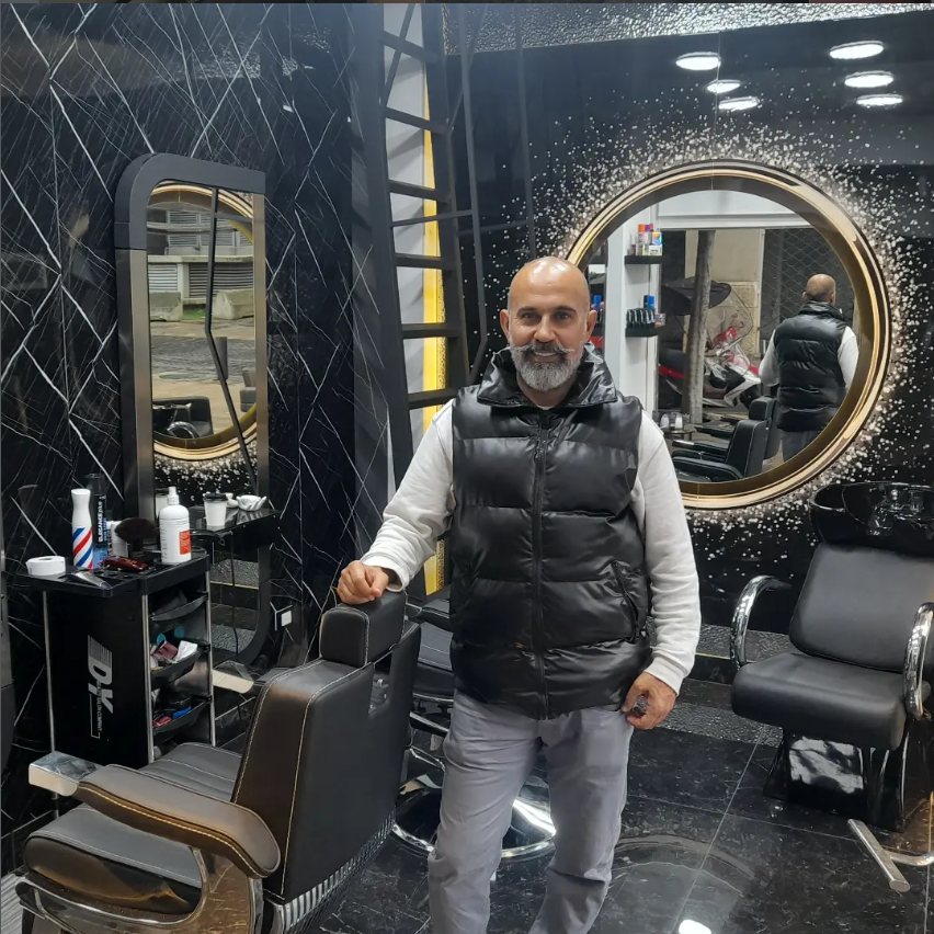 The owner of MANGO Salon standing in the shop, between the barber chairs and the gold-framed mirror